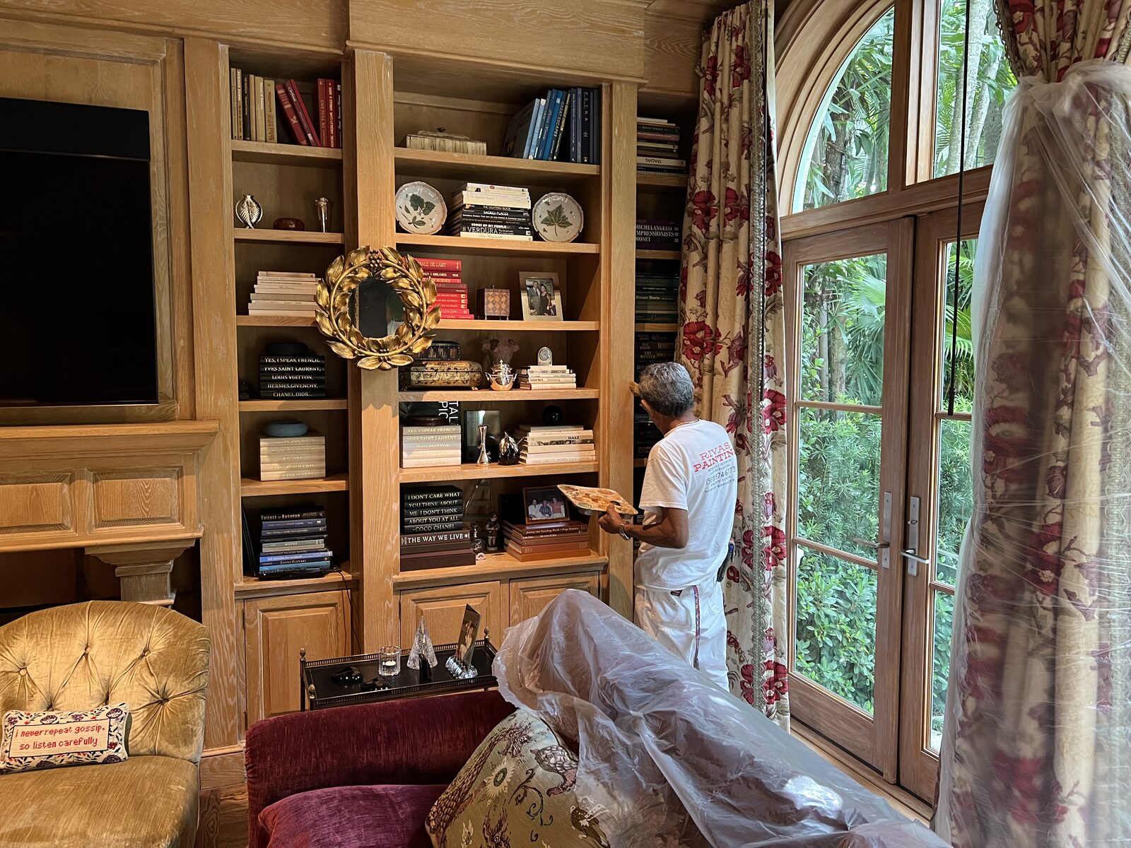 Painter hand-finishing oak library built-ins with furniture wrapped in plastic